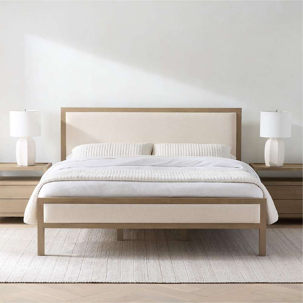 Crate & Barrel Keane Weathered Natural Wood Upholstered Queen Bed is styled in a bright bedroom with matching nightstands, white bedding, and table lamps