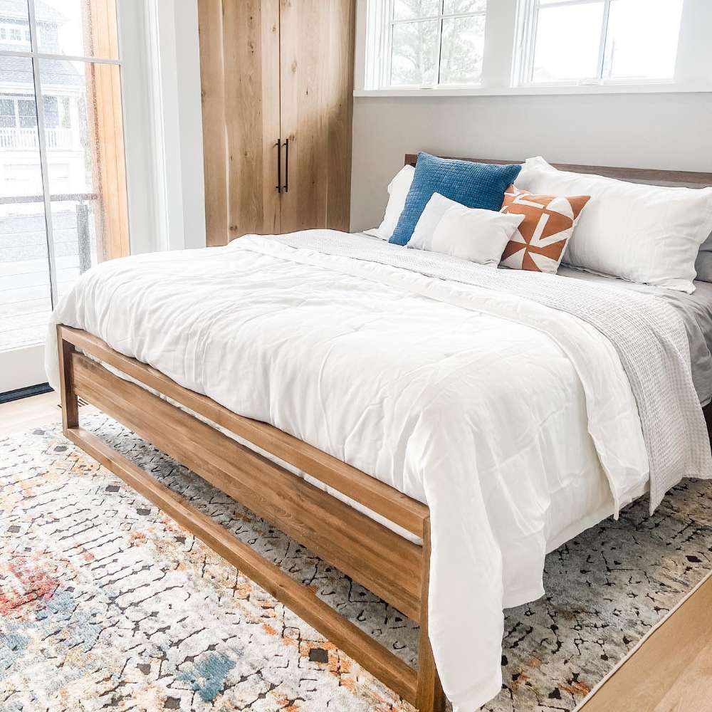 Crate & Barrel Linea Natural Teak Wood Bed in a sunny bedroom with white bedding, colorful pillows, a patterned rug, and wood closet doors