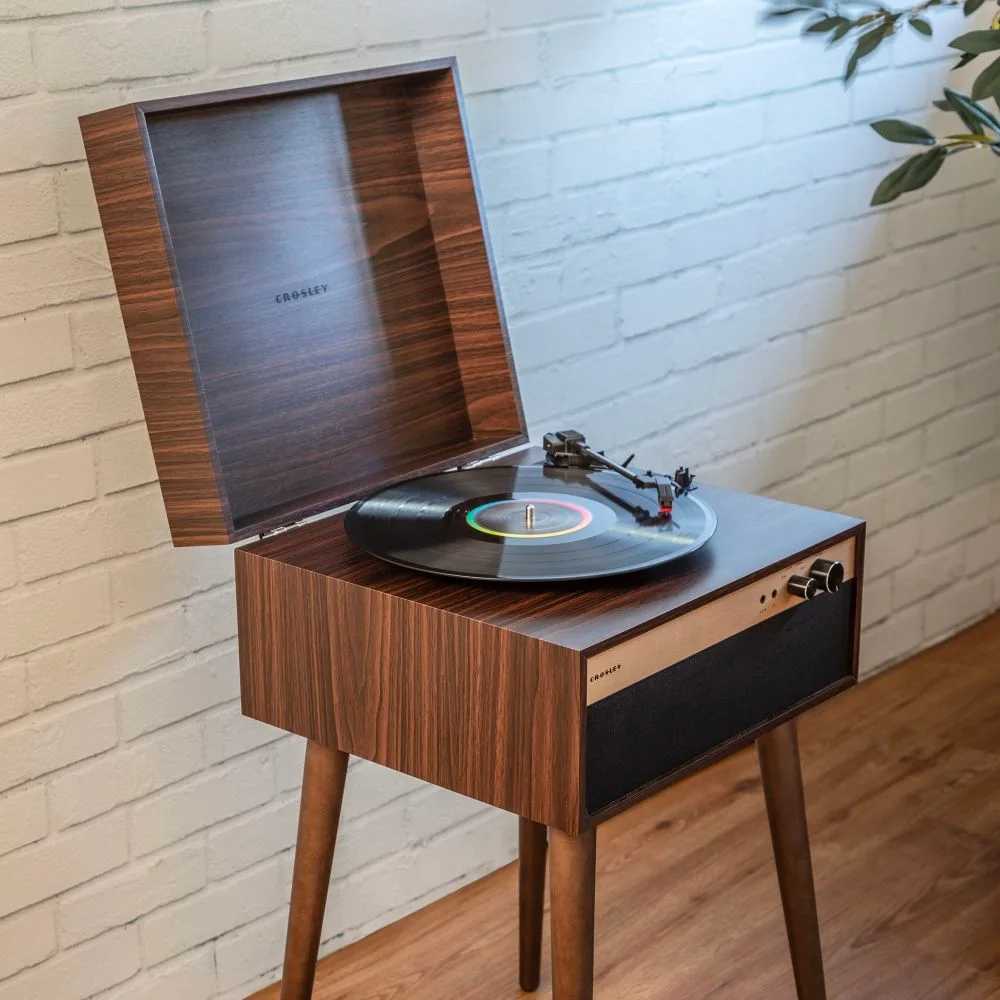 Crosley Jasper Turntable - Walnut is shown playing a vinyl record, standing on wooden legs in a modern, minimalist room