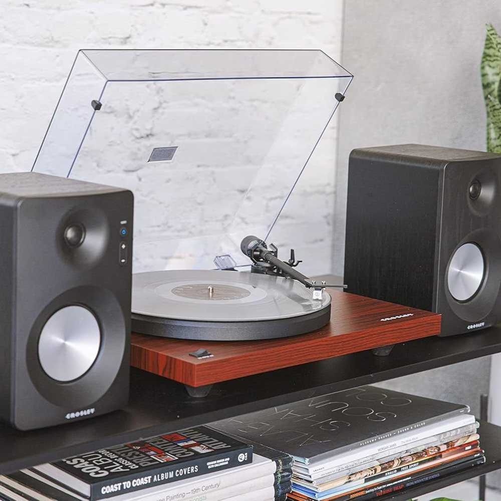 Crosley KT6101 K-Series Turntable System - Mahogany is displayed on a shelf with its dust cover open and vinyl spinning, paired with two stereo speakers