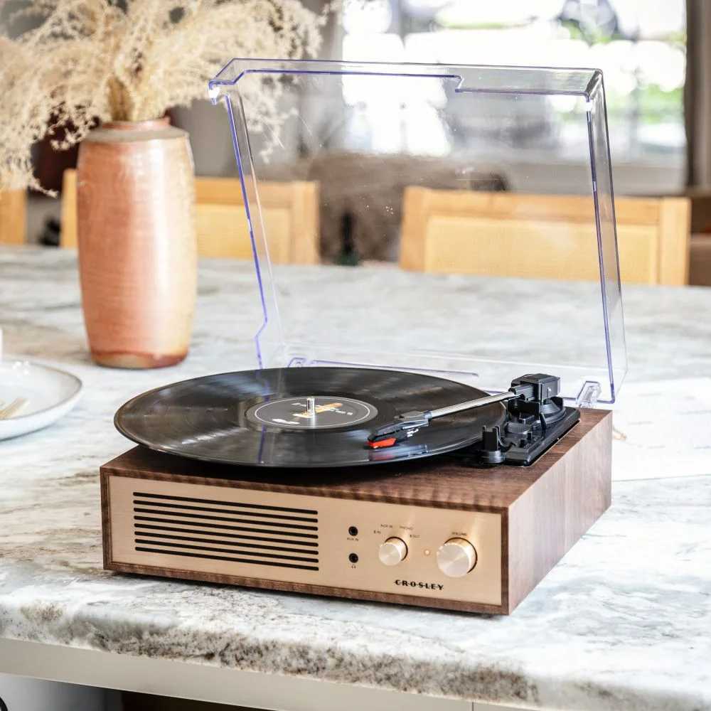 Crosley Miles Record Player - Walnut is showcased on a marble countertop, spinning a vinyl record with its stylish walnut finish and clear protective lid open