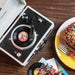 Crosley Mini Cruiser Turntable - White Sand on a wooden table with a colorful vinyl record spinning and a dessert nearby