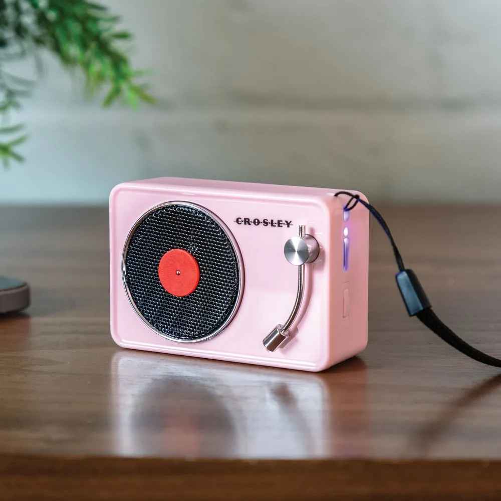 Crosley Mini Turntable Bluetooth Speaker - Pink is a compact speaker shaped like a retro turntable, resting on a wooden table