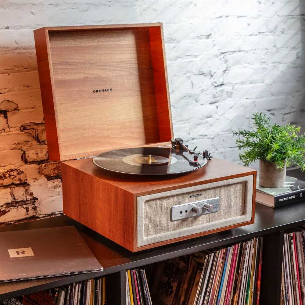 Crosley Rohe Turntable - Natural is set on a bookshelf filled with records, emphasizing its compatibility with vintage and modern interiors