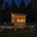 Cube Loft 100 garden room glowing warmly at night with illuminated windows, roof downlights, pathway lights, and two outdoor lounge chairs beside the forest.