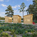 Three Cube Loft 100 garden room units arranged along a landscaped forest path beside a lake, with pale wood exteriors, sloped roofs, pathway lights, and a red Adirondack chair.