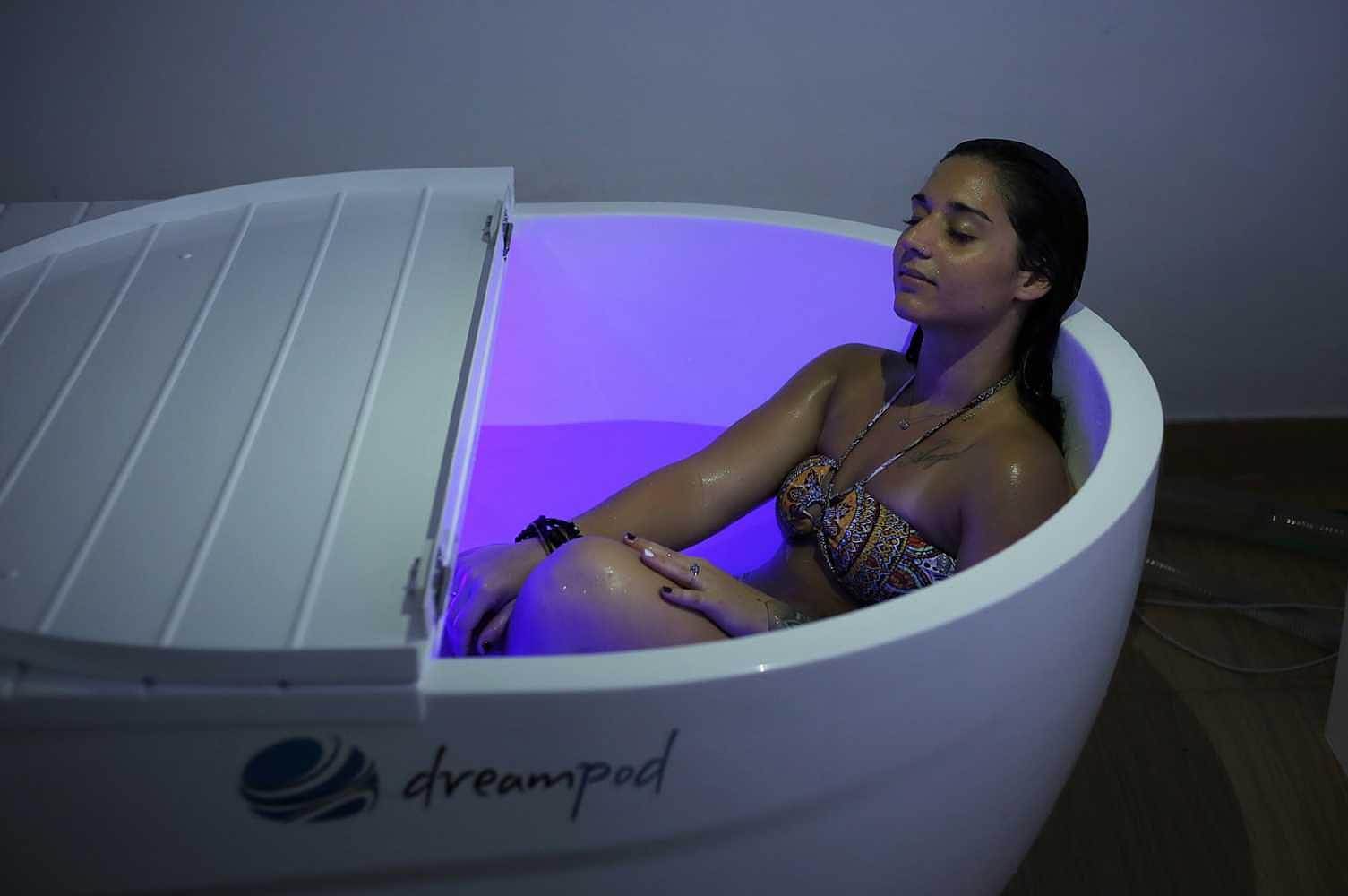 Dreampod Mineral Soaker, with a partially closed lid, features a woman inside enjoying a tranquil float, bathed in soft purple lighting