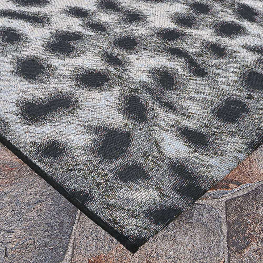 Edge of a Couristan Dolce Indoor/Outdoor Rug - Lynx/Ivory-Charcoal is shown lying on stone tiles, with a fuzzy black and white abstract pattern visible