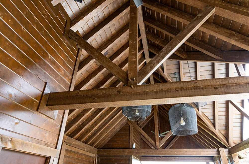 Exposed wooden beams and elegant hanging lights add a rustic yet refined touch to the vaulted ceiling of the Brookside Timber Frame Denalivue Pavilion