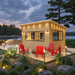 Eze-Breeze Sunroom Cabin illuminated at dusk beside a lake, featuring black-framed windows, five red Adirondack chairs, deck lights, and lush landscaping.