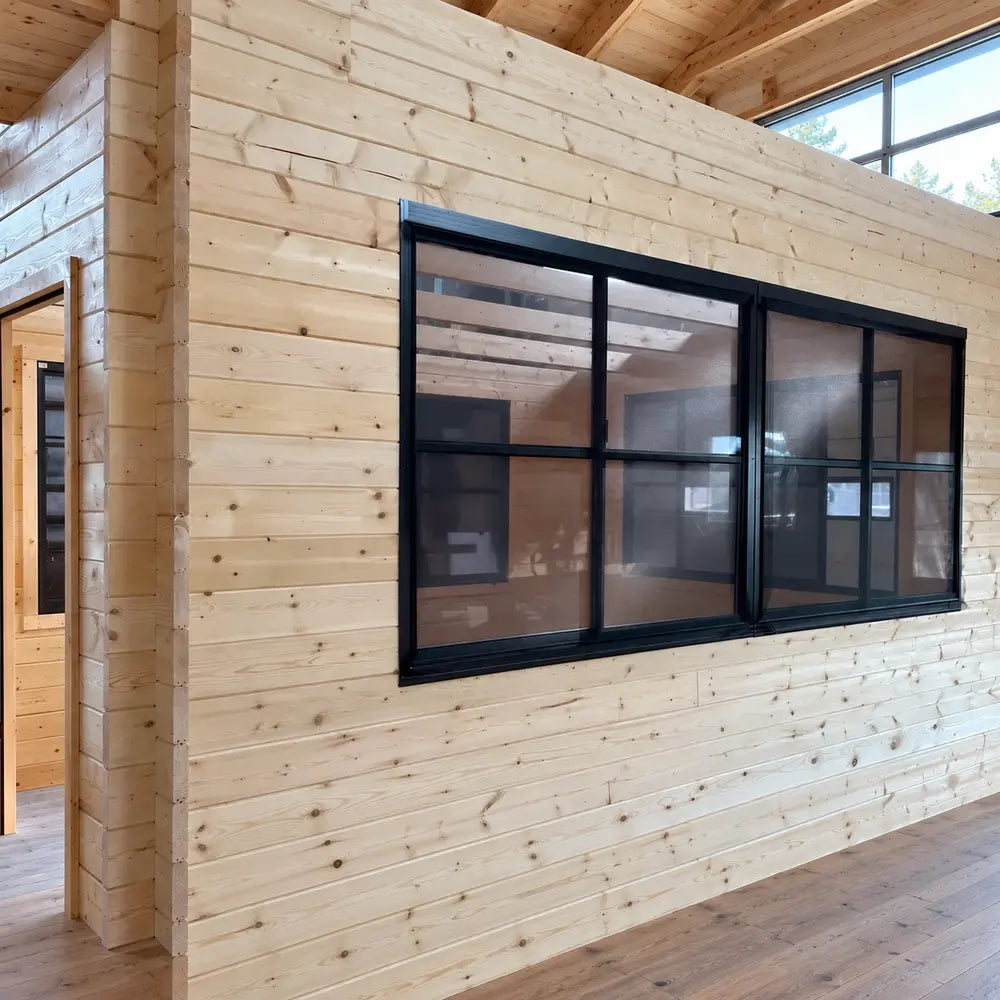 Angled interior close-up of an Eze-Breeze black-framed sliding-window system in a pine wall, with an interlocking timber corner, nearby doorway, and clerestory glazing.