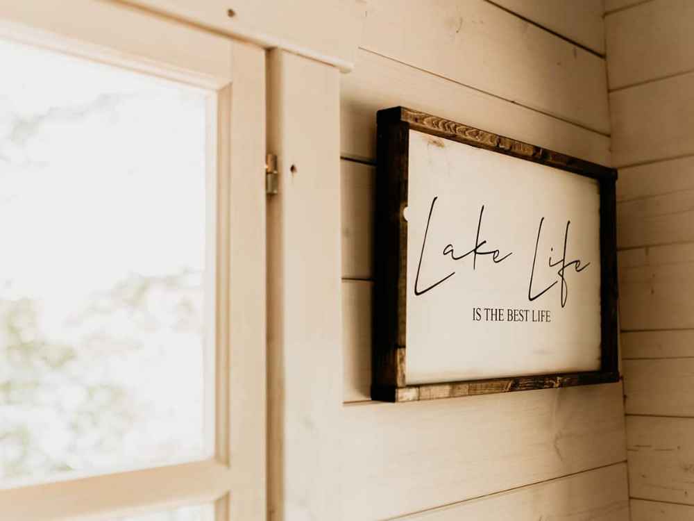 Framed wall sign that reads “Lake Life is the Best Life” next to a wooden-framed window inside the Sawmill Structures Harvest Grand Bungalow 107 Garden Room