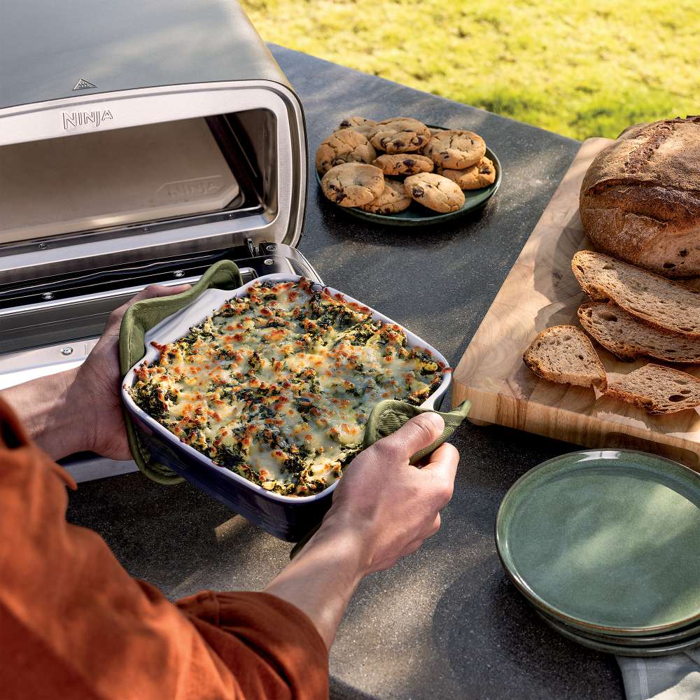 Fresh bread, cookies, and a baked casserole sit beside Ninja Artisan 5-in-1 Portable Electric Pizza & Outdoor Oven outdoors