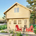 Front-facing view of the Sawmill Structures Grand Loft 107 Deluxe DD Garden Room shows its wood construction, large loft window, and red chairs arranged on the patio