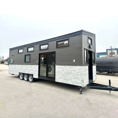 Full side view of Sawmill Structures Wanderlove Tiny Home shows its triple-axle base, black top siding, and stone-textured bottom half