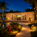 Game Room 70 glowing brightly at night as family and friends watch sports inside, framed by illuminated palms, tropical plants, and Florida lagoon reflections.
