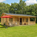 GardenHouse24 USA Barrit 70 Garden Room stands in a grassy yard with a red umbrella and patio seating on the porch