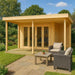 GardenHouse24 USA Keno 70 Garden Room is set up in a lush garden, featuring large glass doors and a covered porch with wicker furniture