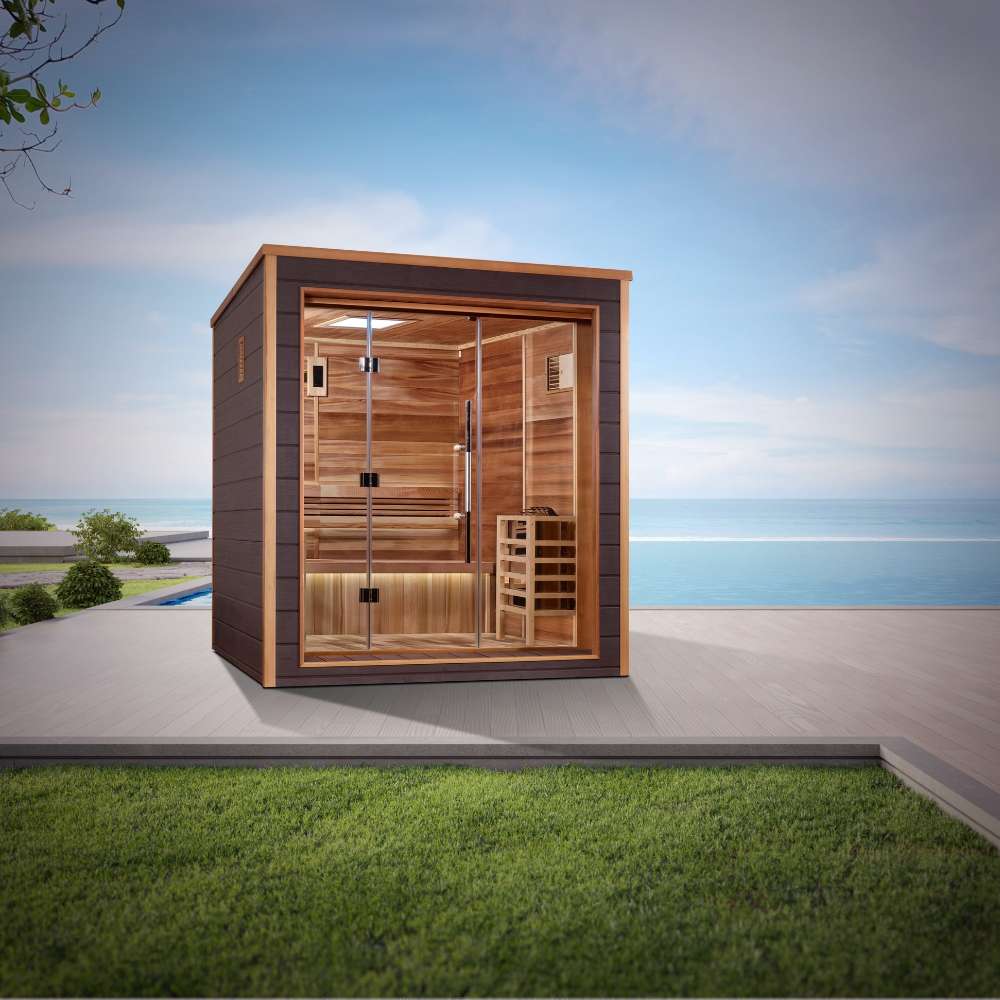 Golden Designs Drammen 3 Person Outdoor-Indoor Traditional Sauna sits on a gray patio beside green grass with the sea and a blue sky behind it