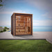Golden Designs Drammen 3 Person Outdoor-Indoor Traditional Sauna sits on a gray patio beside green grass with the sea and a blue sky behind it