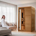 Golden Designs Dynamic Lucca Elite 2 Person Ultra Low EMF Far Infrared Sauna stands in a bright living room beside a sofa where a woman in a white robe relaxes