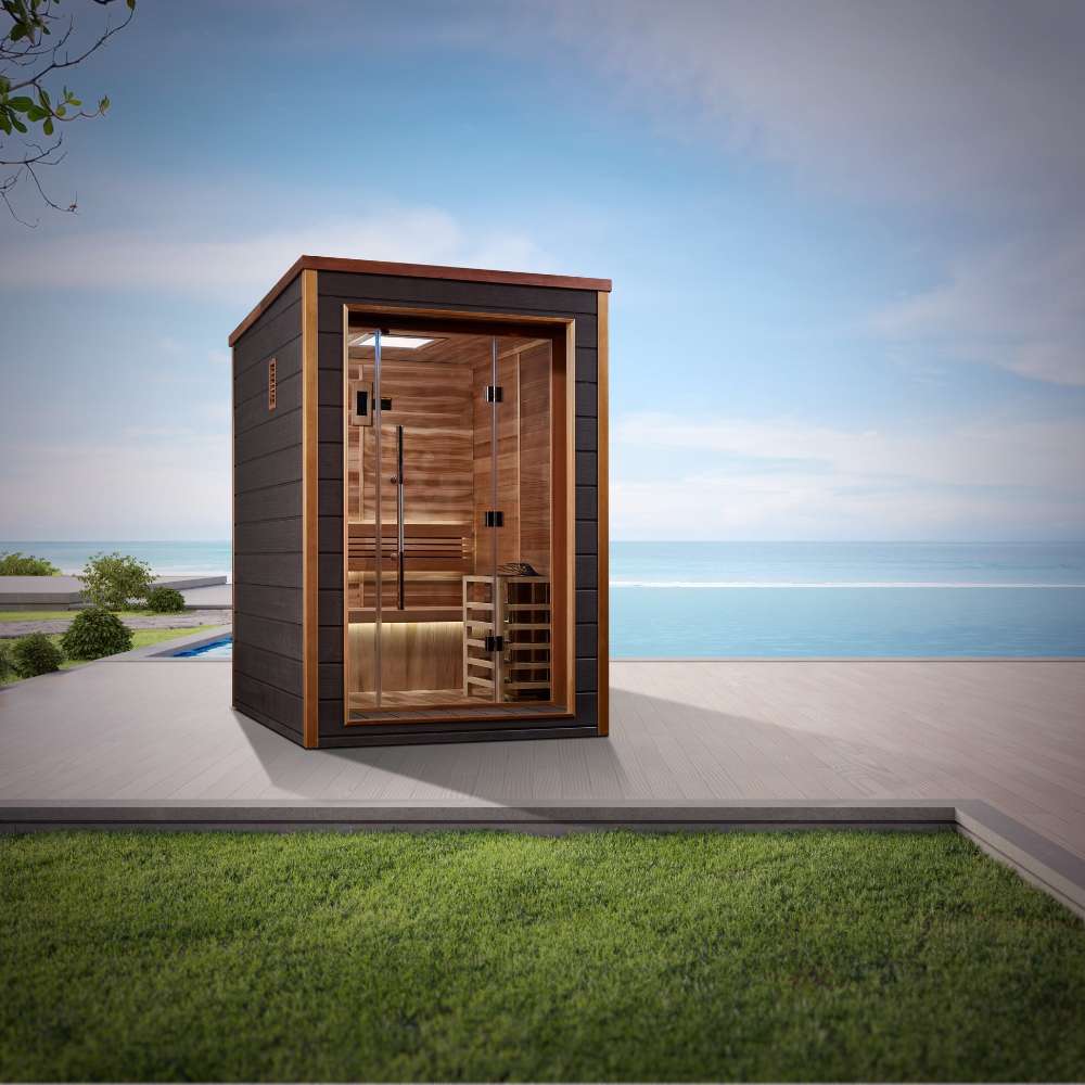 Golden Designs Narvik 2 Person Outdoor-Indoor Traditional Sauna stands on a stone patio beside a lawn with the sea and sky behind it