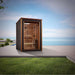 Golden Designs Narvik 2 Person Outdoor-Indoor Traditional Sauna stands on a stone patio beside a lawn with the sea and sky behind it