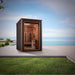 Golden Designs Nora 2 Person Outdoor-Indoor PureTech Hybrid Full Spectrum Sauna stands on a stone patio beside a lawn with the sea and sky behind it