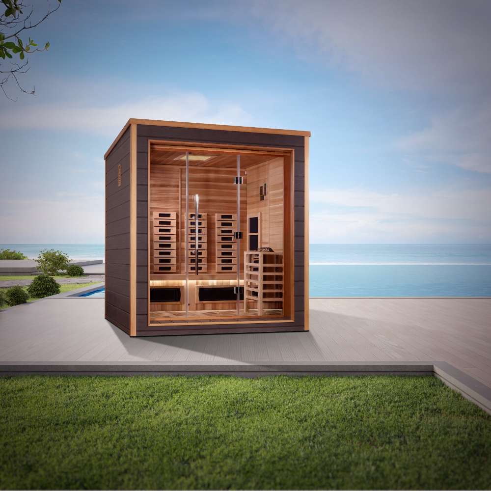 Golden Designs Visby 3 Person Outdoor-Indoor PureTech Hybrid Full Spectrum Sauna sits on a gray patio beside green grass with the sea and a blue sky behind it