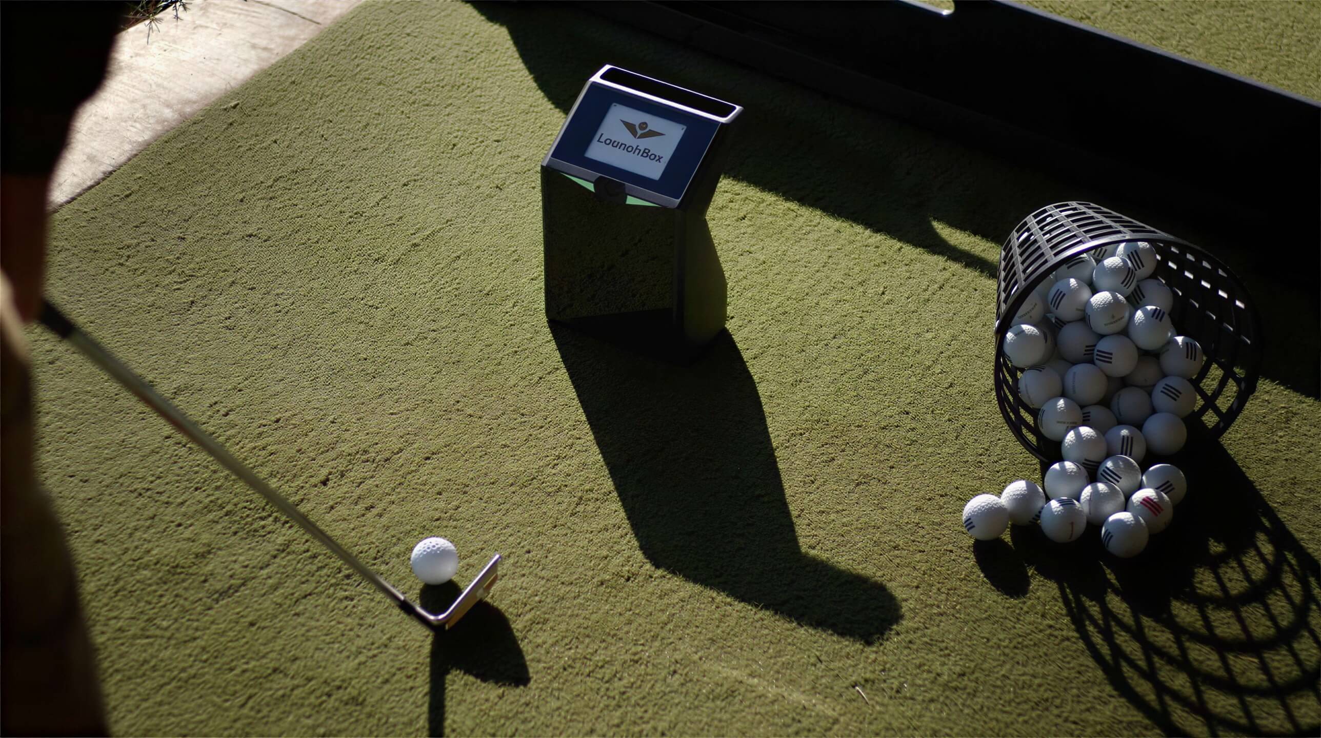 Golf ball sits on indoor turf next to a driver, with TruGolf LaunchBox Golf Launch Monitor w/ E6 CONNECT Home positioned nearby to measure the shot