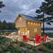 Angled view of the GrandView 150 wooden cabin with a metal roof, illuminated deck, red Adirondack chairs and a lake behind it.