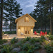 Front view of the two-story GrandView 150 cabin glowing warmly in a forest clearing with red Adirondack chairs and pathway lights.