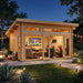 Twilight view of a timber cabin with a lively interior sports watch party. Through large glass doors, people are cheering at a television. Outside is a lit stone patio with a fire pit and trees under a starry sky.