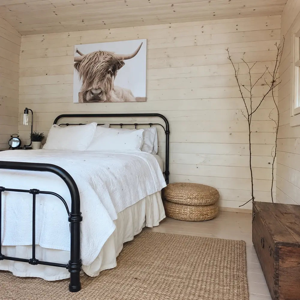 Cozy Harvest Grand Bungalow bedroom with a black metal bed, white bedding, woven poufs and a wall-mounted Highland cow portrait.