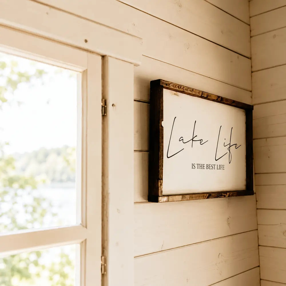 Framed “Lake Life Is the Best Life” wall sign beside a sunlit wooden window inside the Harvest Grand Bungalow.