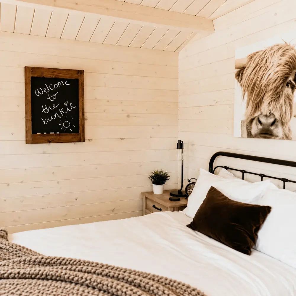 Harvest Grand Bungalow bedroom featuring a “welcome to the bunkie” chalkboard, Highland cow artwork and rustic bedside décor.