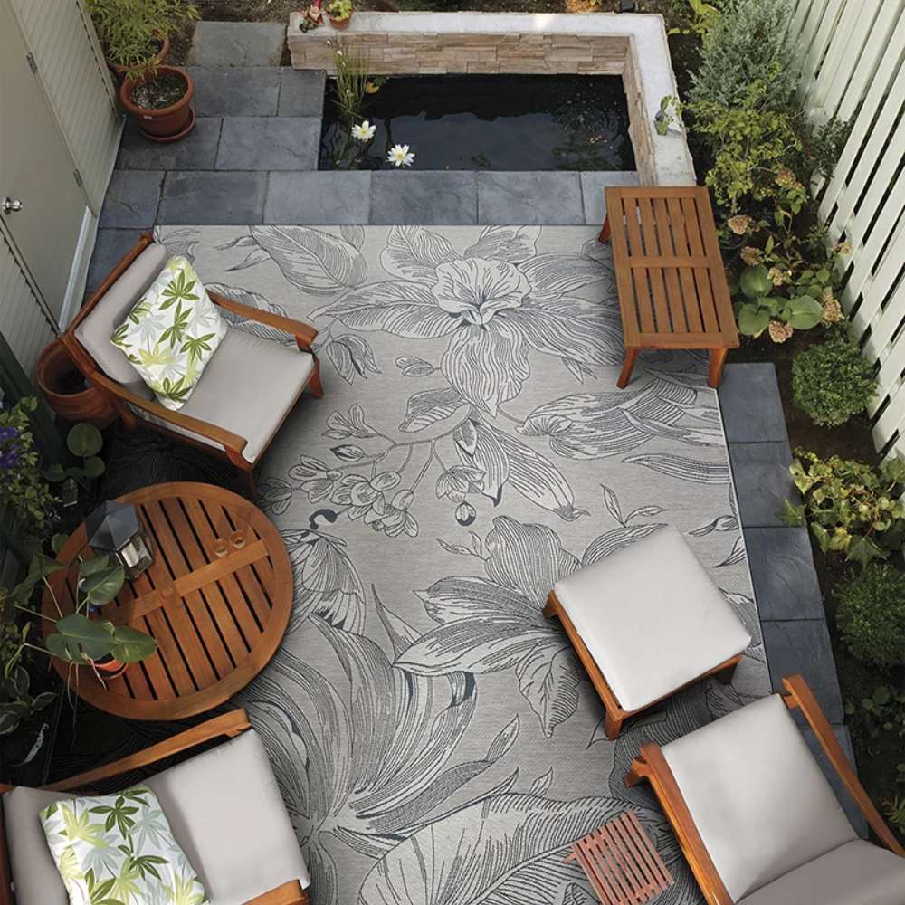 In a cozy patio setup, Couristan Dolce Indoor/Outdoor Rug - Flora/Argento anchors wooden furniture and surrounds a small pond