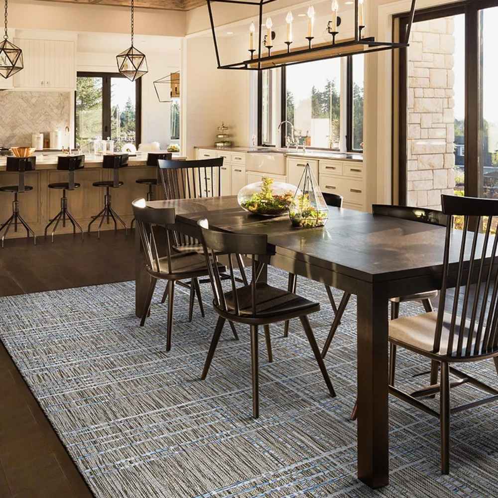 In a modern dining room, the Couristan Charm Indoor/Outdoor Rug - Ohe/Sand-Ivory lies under a dark wood table, adding subtle elegance with its muted grid design