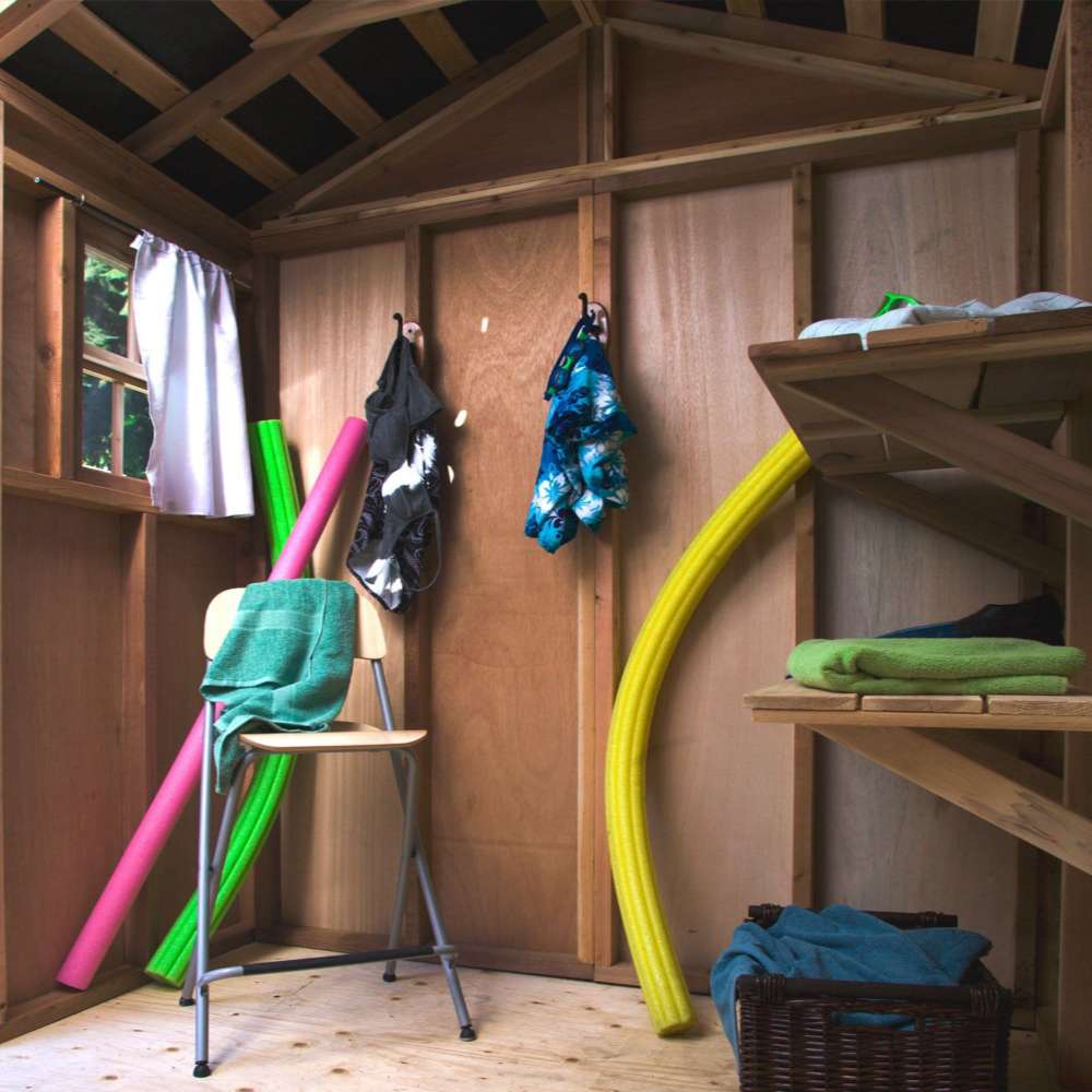 Inside a wooden storage shed, colorful pool noodles, towels, and hanging swimwear are organized neatly on wall-mounted Outdoor Living Today 8ft Western Red Cedar Shed Shelves (Set of 3)