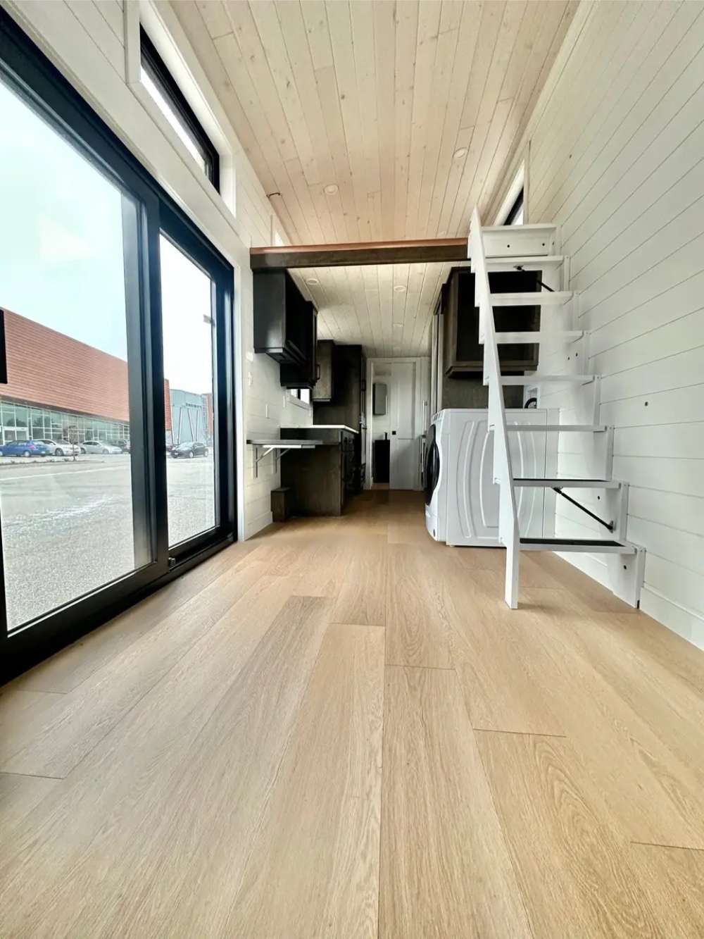 Inside Sawmill Structures Wanderlove Tiny Home, there’s a bright open hallway with wood flooring, white shiplap walls, and a stairway leading to a loft