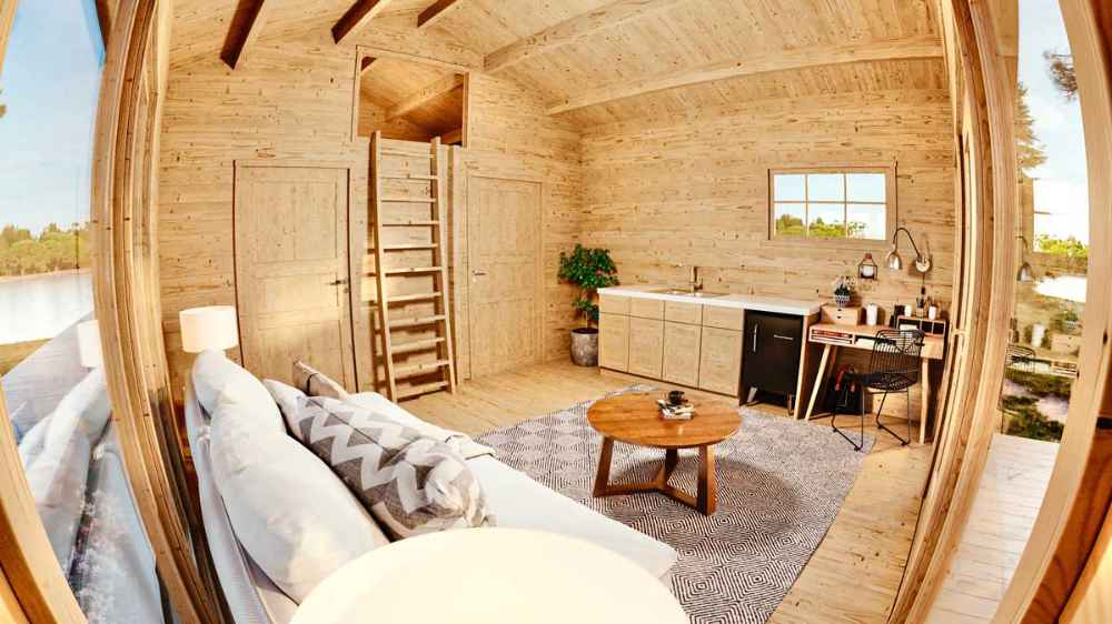 Inside the Sawmill Structures Edwin Lake House 70MM Garden Room, a cozy living space features a couch, rug, small round table, and a compact kitchen along one wall