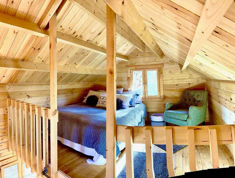 Inside the Sawmill Structures The Jumbo Grand Loft 130 SD Garden Room, the lofted sleeping space includes a cozy bed, “Get Cozy” pillows, and a reading nook with a green chair