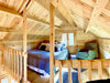 Inside the Sawmill Structures The Jumbo Grand Loft 130 SD Garden Room, the lofted sleeping space includes a cozy bed, “Get Cozy” pillows, and a reading nook with a green chair