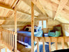 Inside the Sawmill Structures The Jumbo Grand Loft 130 SD Garden Room, the lofted sleeping space includes a cozy bed, “Get Cozy” pillows, and a reading nook with a green chair