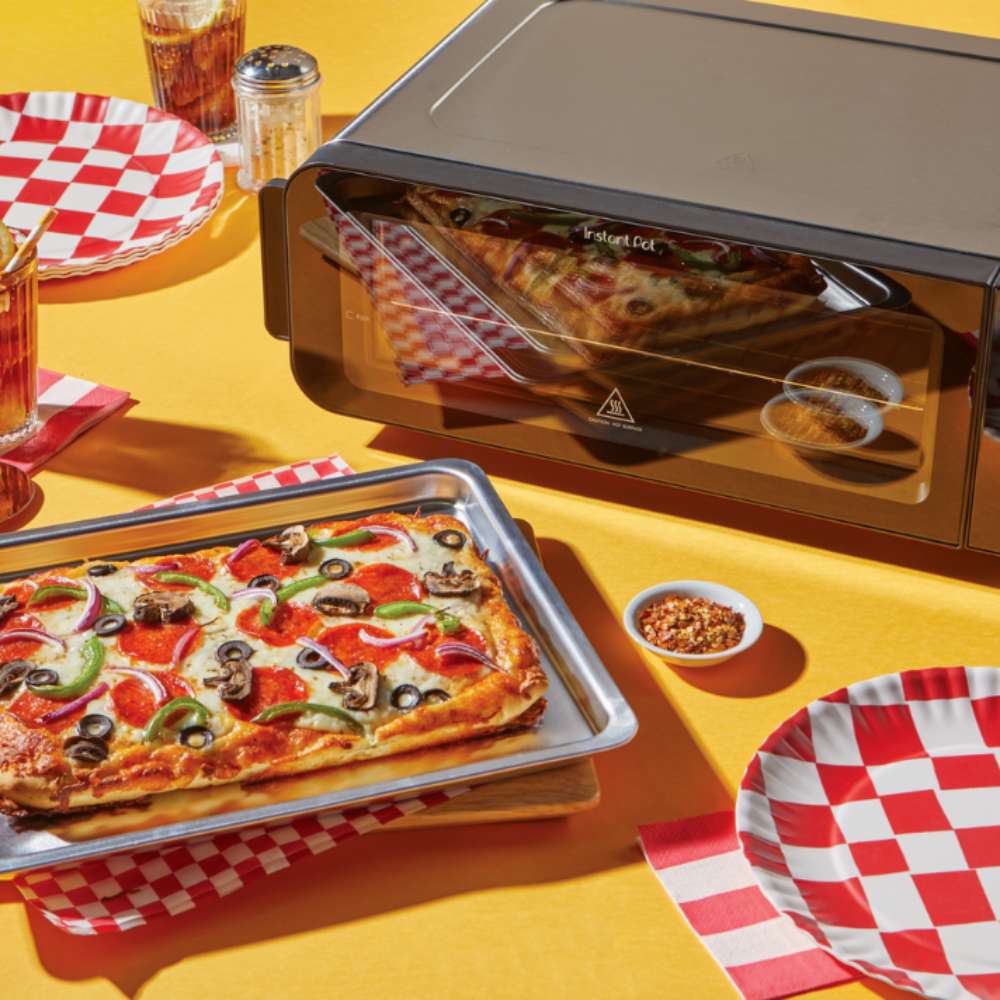 Instant Pot® InstantHeat™ Pizza Oven sits on a yellow table beside a rectangular pizza, checkered plates, and seasonings