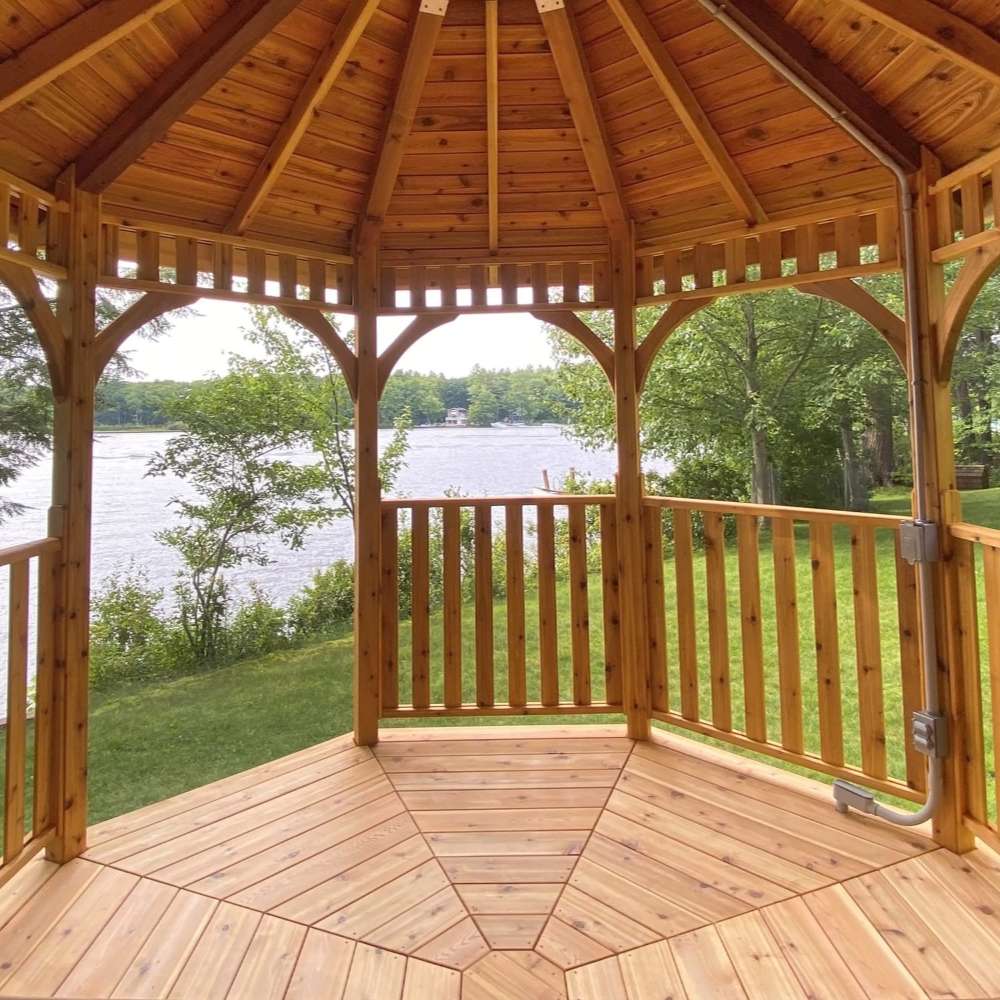 Interior view of Outdoor Living Today Western Red Cedar Octagon Bayside Gazebo shows a wooden floor and open sides overlooking a calm lakeside