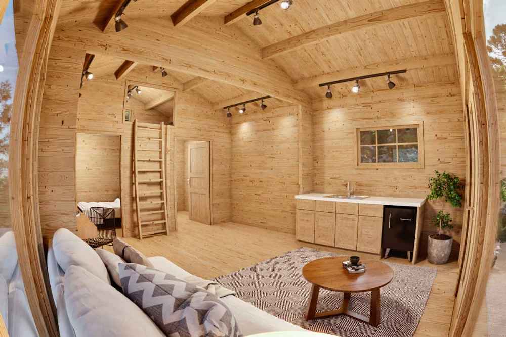 Interior of the Sawmill Structures Edwin Lake House PLUS 70MM Garden Room features an open layout with a kitchenette, round coffee table, loft ladder, and exposed wooden beams