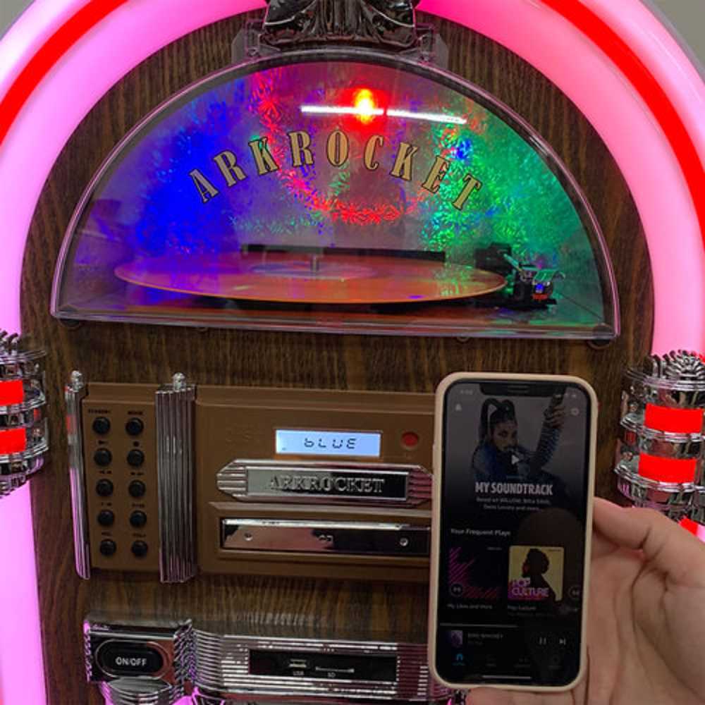 In the image, a person holds a phone in front of Arkrocket Taurus II Jukebox with Stand - Dark Oak 2024 New Upgrade showing Bluetooth connectivity