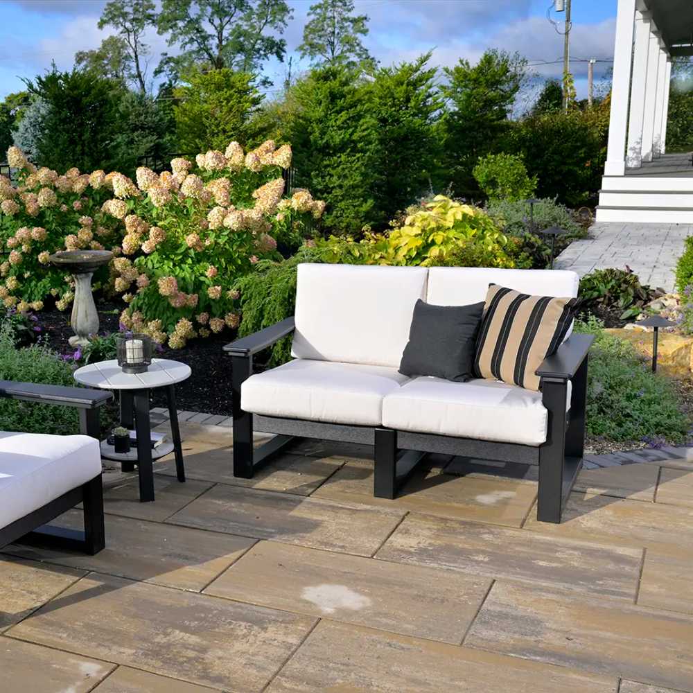 In this garden setting, the LuxCraft Lanai Deep Seating Loveseat showcases white cushions with a black frame, paired with decorative throw pillows