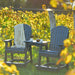 In this image, two Adirondack-style chairs sit on grass next to a vineyard, connected by a LuxCraft Balcony Tete-a-Tete Table that holds coffee cups and a snack bowl